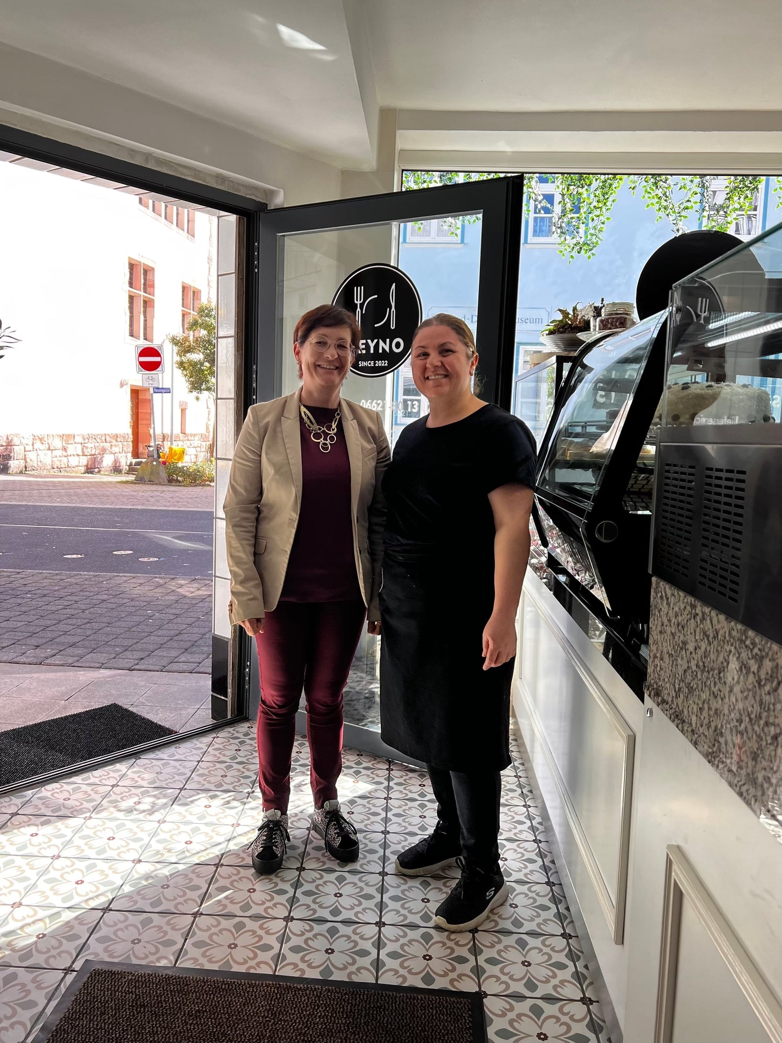 Anke Hofmann und Zeynep Kaya stehen lächelnd im Eingang des Zeyno Cafés. Hinter ihnen ist das Café-Logo und der Slogan "Since 2022" zu sehen.