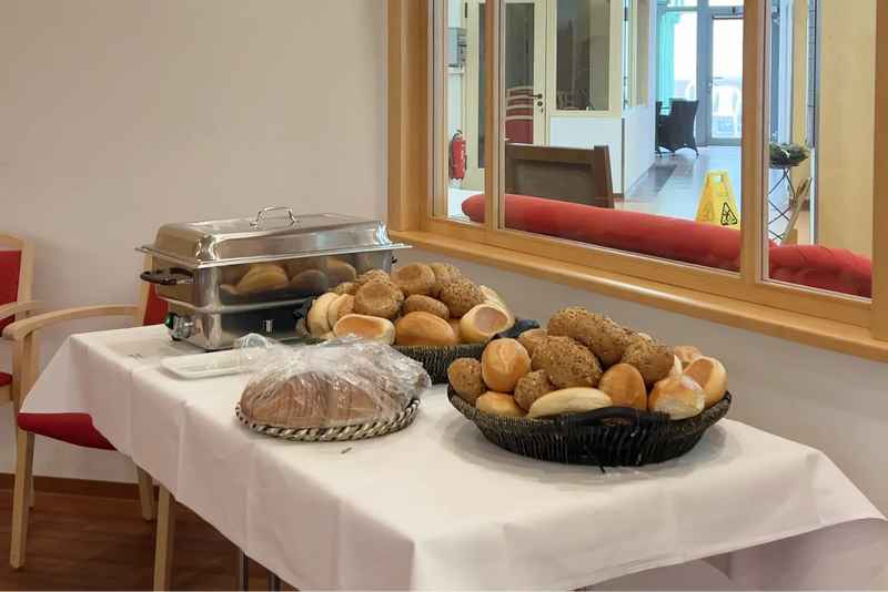Ein Buffettisch mit weißem Tischtuch, auf dem zwei große Körbe mit verschiedenen frischen Brötchen (Weizen und Körner), ein verpacktes Brot und ein silberner Chafing-Dish-Warmhaltebehälter stehen.