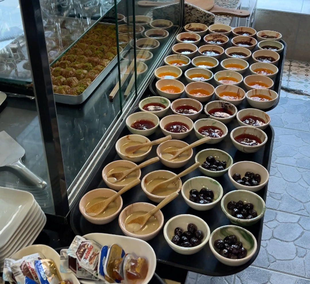 Ein Tisch mit zahlreichen kleinen Schälchen, die verschiedene Marmeladen und Oliven enthalten. Im Hintergrund ist eine Glasvitrine mit weiteren Leckereien zu sehen.