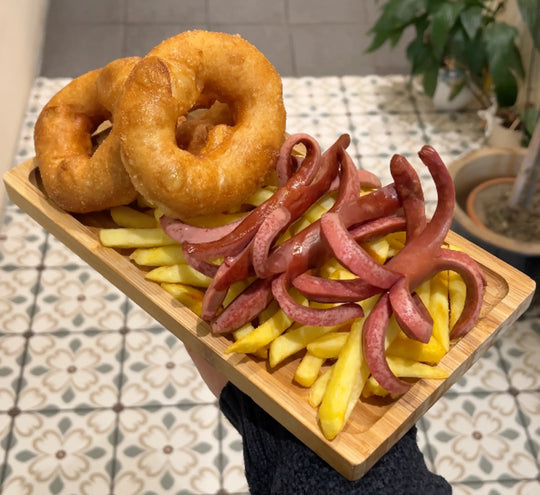 Ein Teller mit Pommes frites, zwei goldbraun frittierten Pişi (türkische "Bagels", ähnlich wie Churros) und aufgeschnittenen Würstchen, die wie kleine Kraken aussehen. Im Hintergrund ist ein gemusterter Fliesenboden zu sehen.